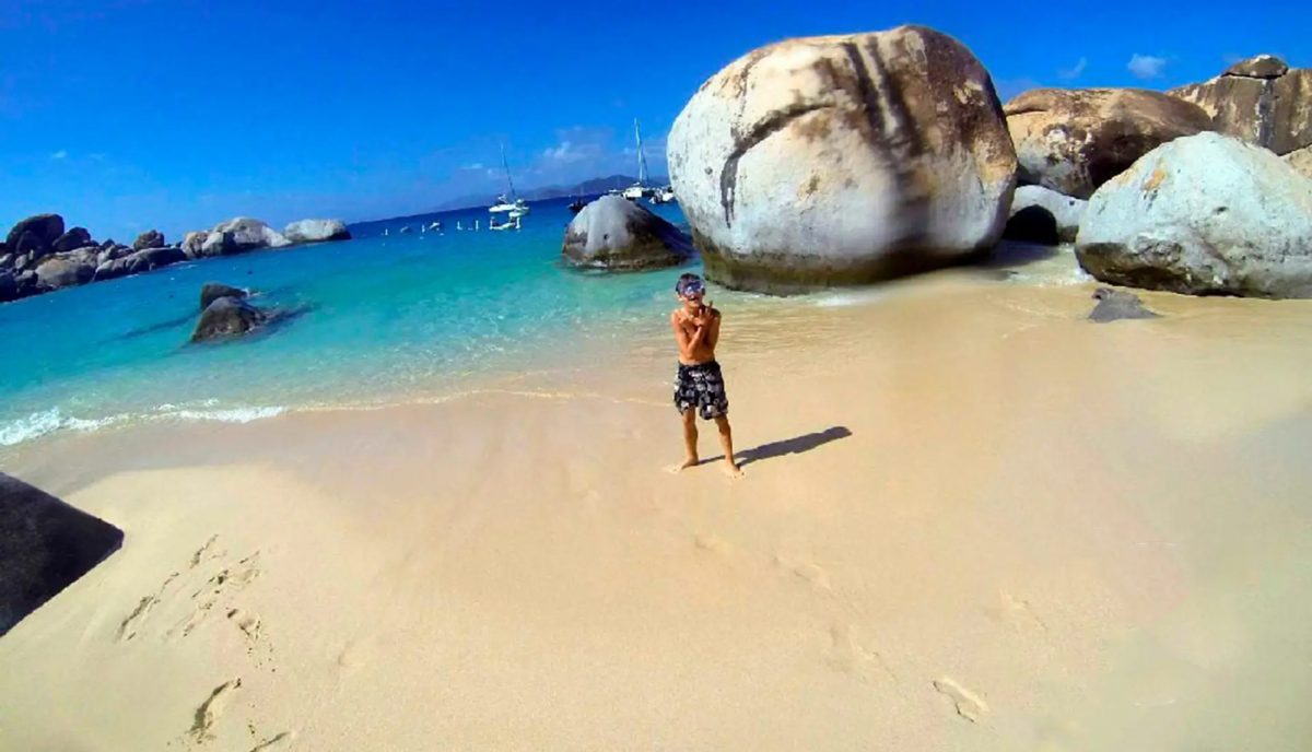 Boy standing on beach at The Baths Virgin Gorda with massive granite boulders and turquoise water with charter yachts anchored in background
